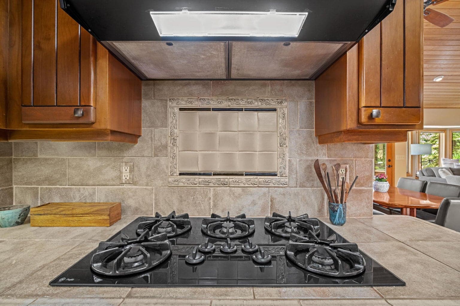 Gas cooktop and range/hood above, neutral tiled counter tops that cover the entire back splash.