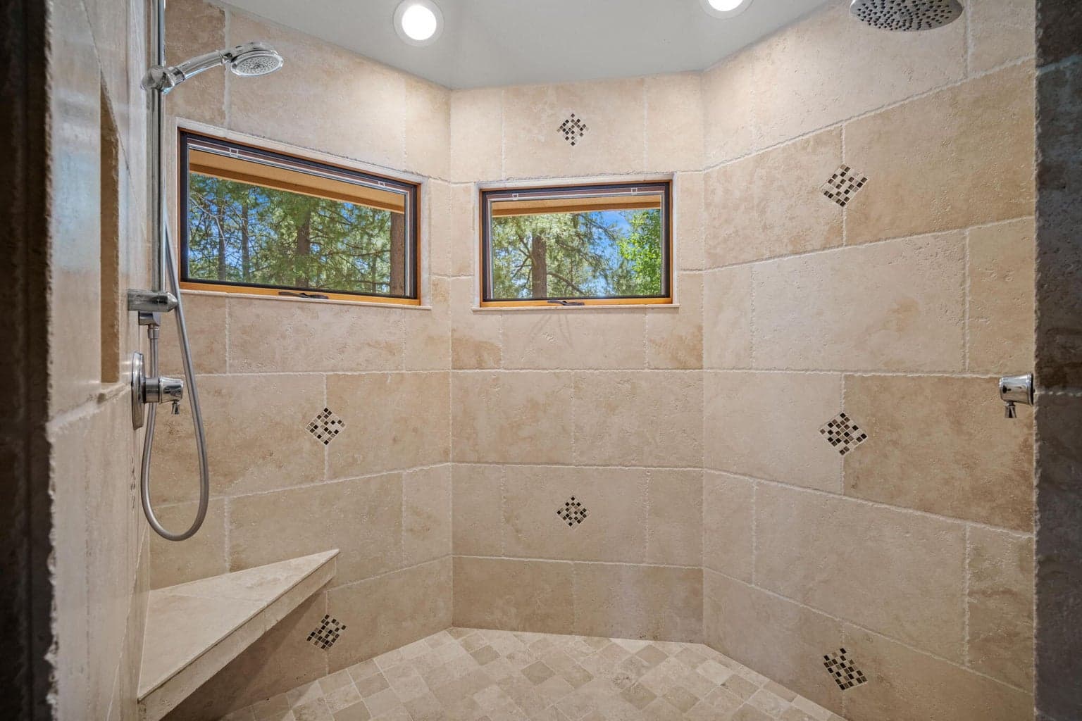 Walk-in travertine shower in the primary suite - with 2 shower heads, built in bench and windows to enjoy a glimpse of the trees outside.