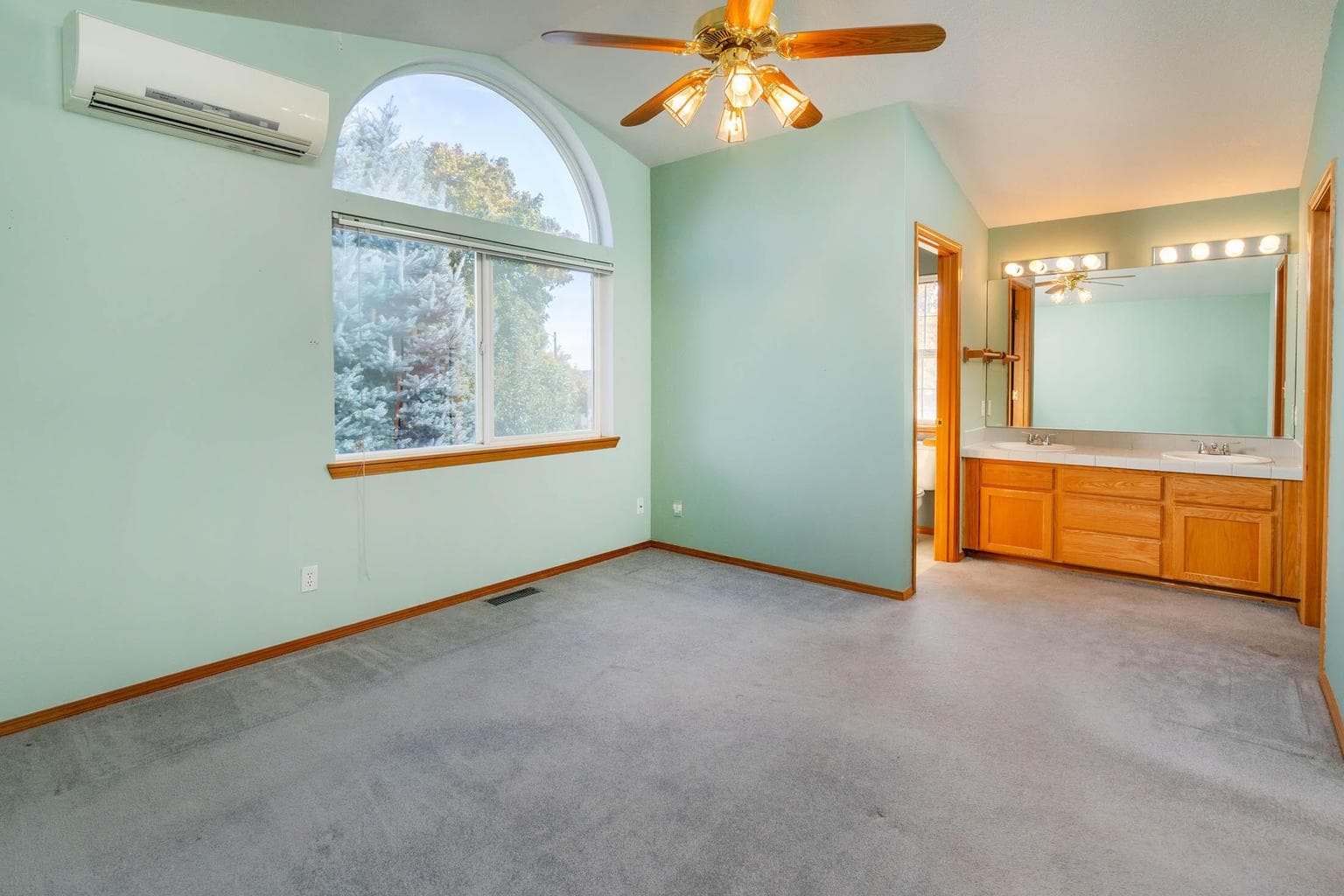 Vaulted primary bedroom w/mini split, ceiling fan, and views out back of home.