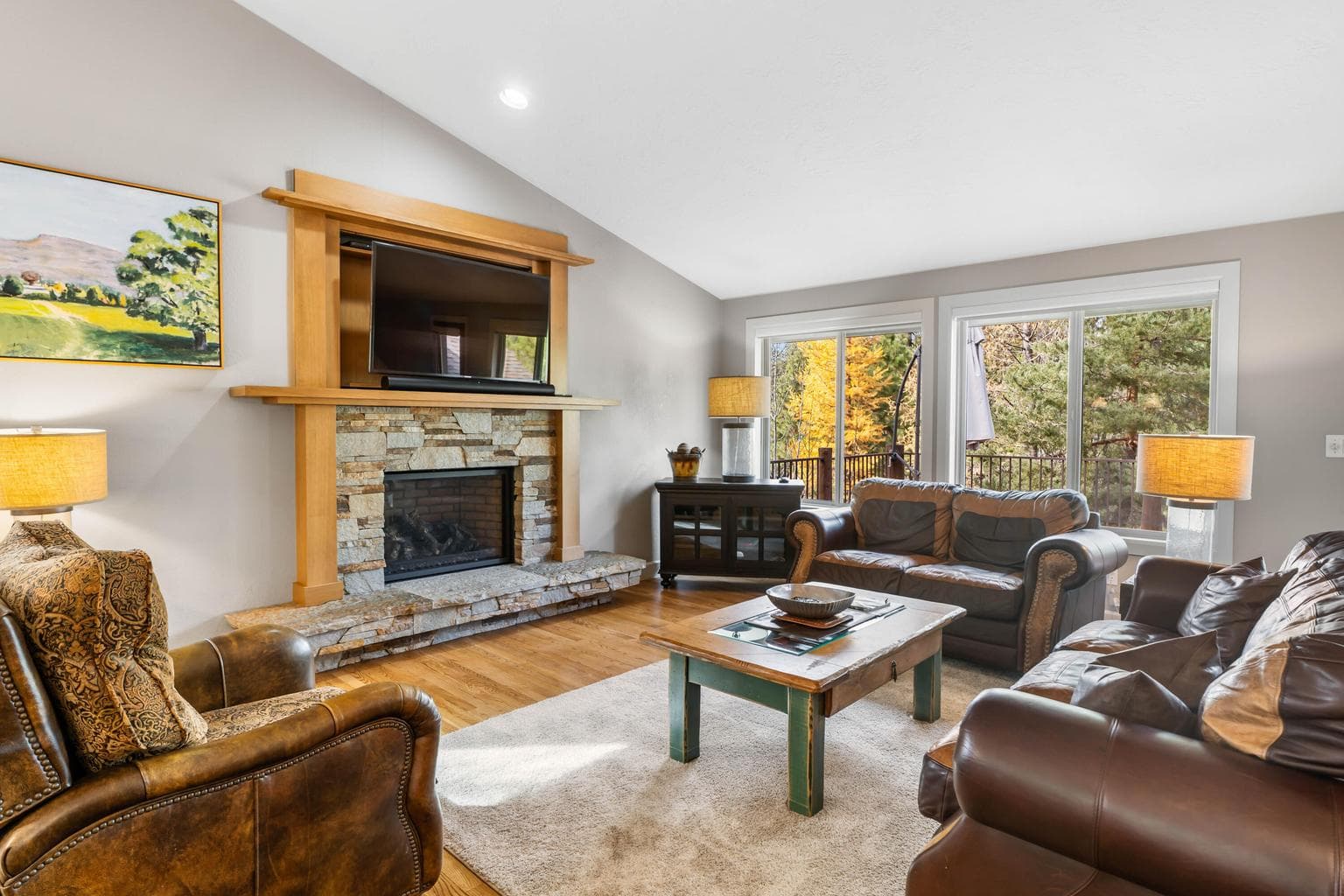 This bright and welcoming great room features a stunning stone fireplace framed in natural wood, rich hardwood floors, and comfortable leather seating. Large windows fill the space with natural light - perfect for enjoying the forest views.