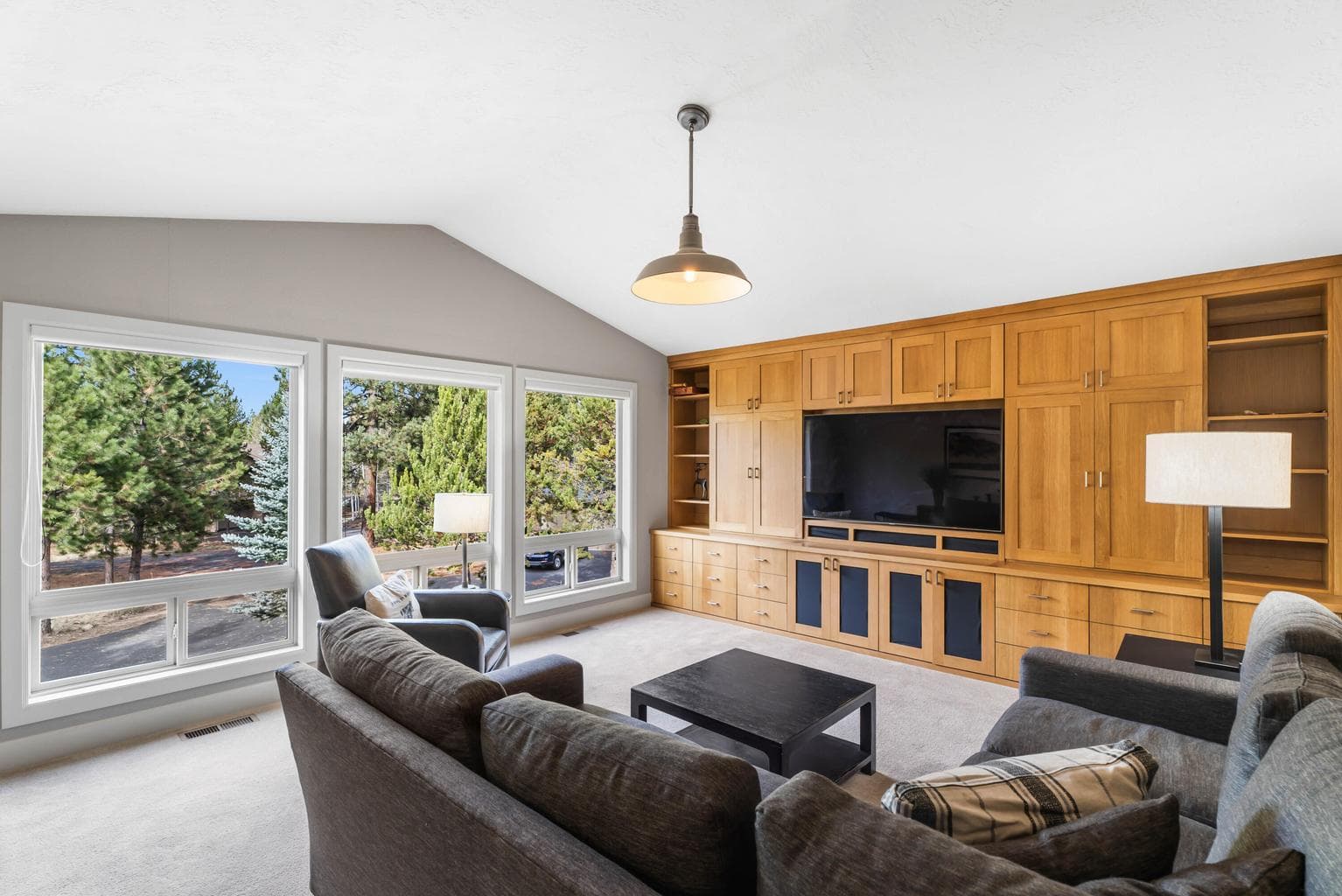 This bright upper-level media room features expansive windows framing treetop views and a wall of custom built-ins for ample storage and display. With vaulted ceilings and cozy seating, it’s the perfect space for movie nights or relaxing in natural light.