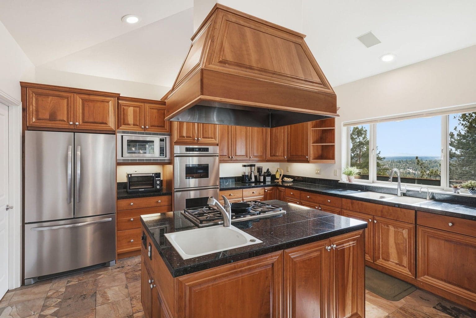 A well-appointed chef’s kitchen with custom cherry hood detail, Dacor appliances, gas cooktop, prep sink, walk-in pantry, and abundant counter space, highlighted by a large window capturing scenic views from the sink.