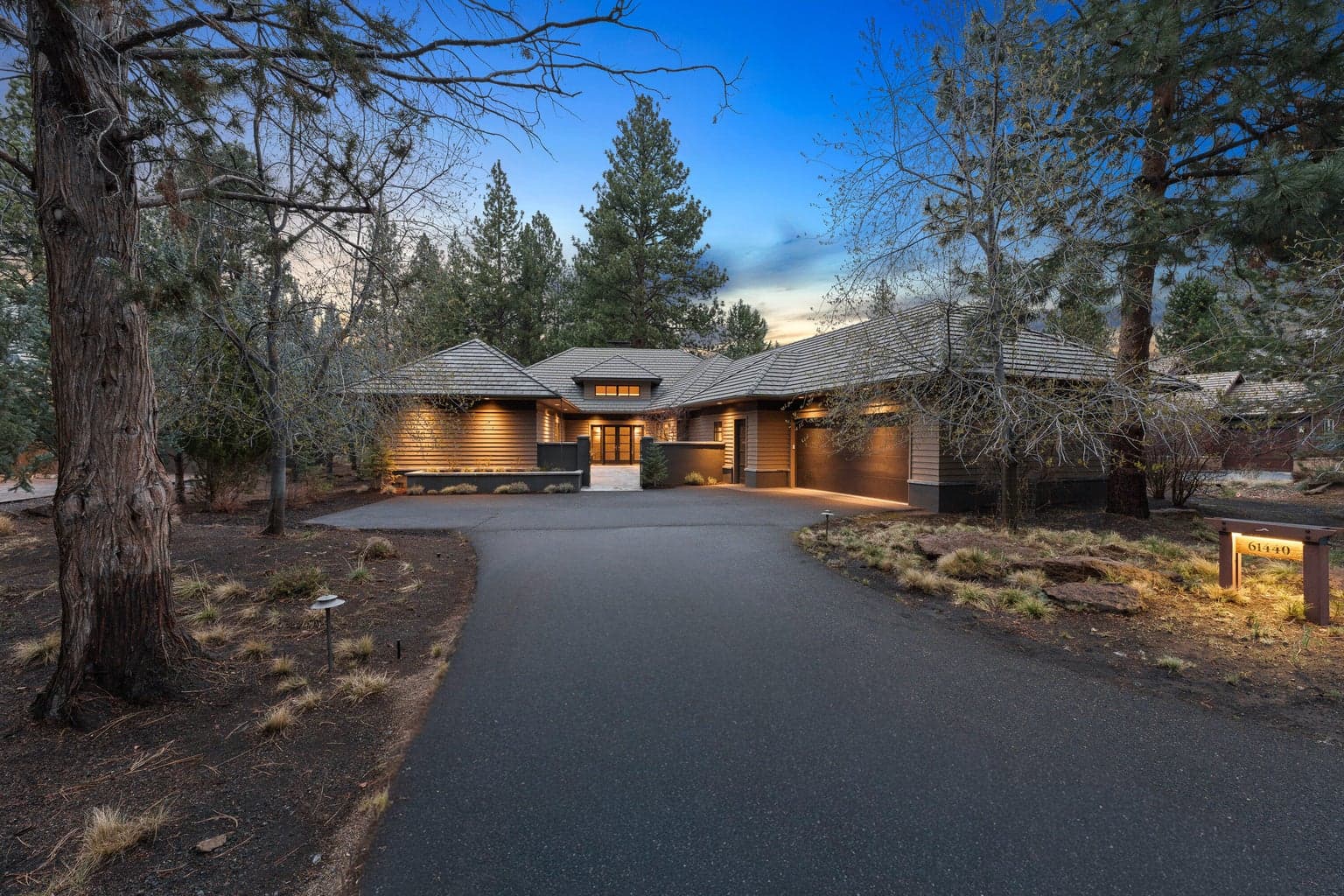 Dusk exterior highlighting the home’s warm, inviting presence, softly illuminated against a backdrop of mature Ponderosa pines. The gently curved driveway leads to a private courtyard entry, creating a sense of arrival that feels both welcoming and refined. Thoughtful exterior lighting and the home’s natural palette enhance its connection to the surrounding landscape, offering a peaceful and elevated setting within the Broken Top community.