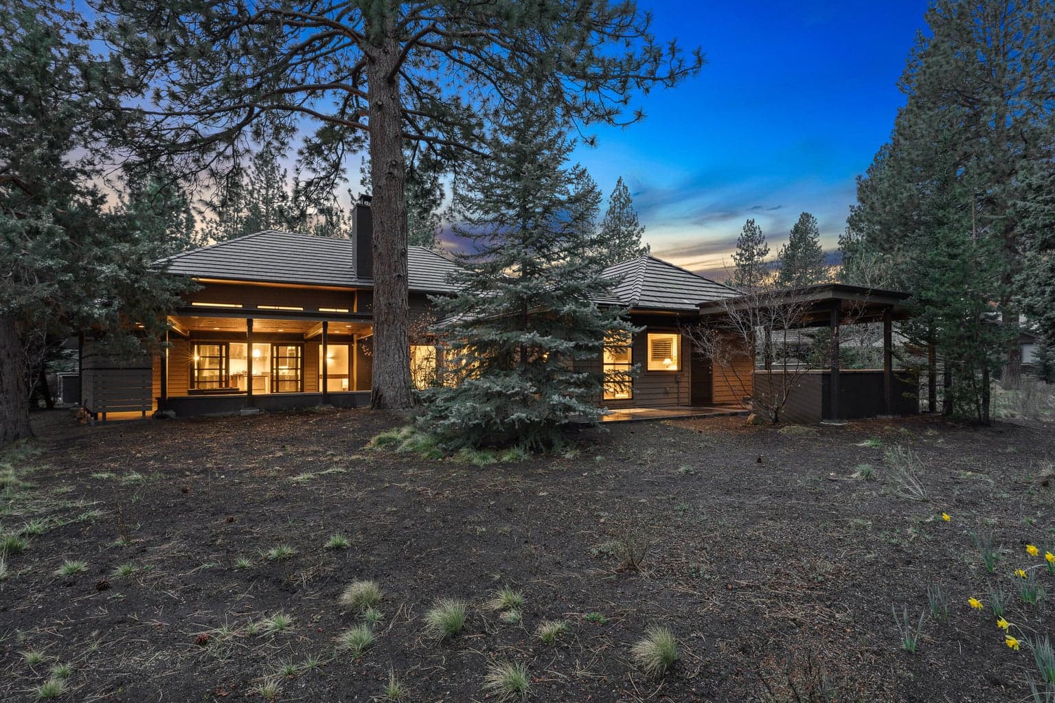 Rear exterior at dusk showcasing the home’s seamless connection to its natural surroundings, set among mature Ponderosa pines with a peaceful, woodsy backdrop. Expansive windows bring a warm glow from within, highlighting the home’s thoughtfully designed living spaces and indoor-outdoor flow. Positioned along the Broken Top golf course, the setting offers a rare combination of privacy, space, and a true sense of retreat.