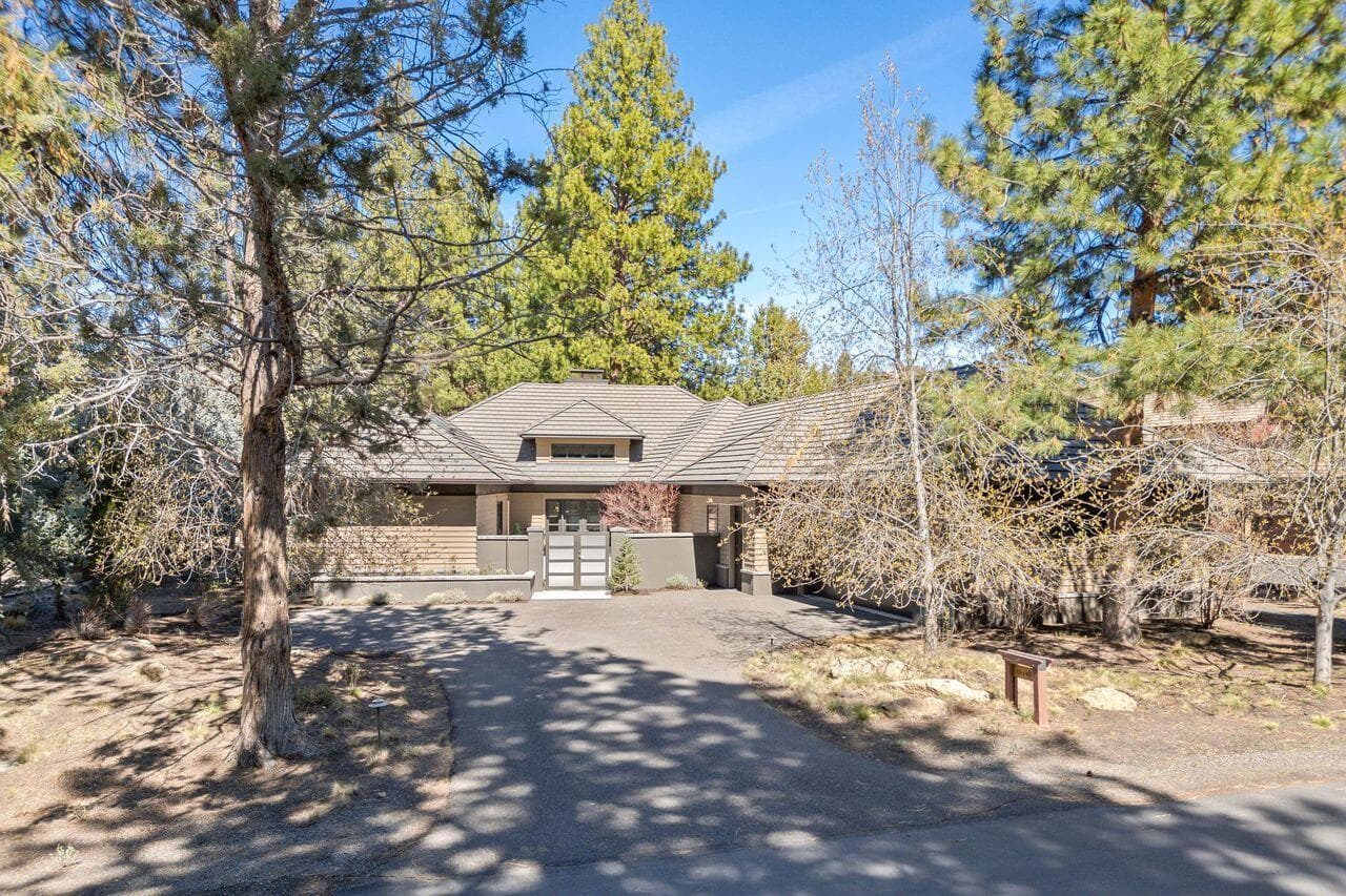 Front exterior showcasing the home’s understated architectural presence, set among mature Ponderosa pines within the gated Broken Top community. A gently curved driveway leads to a private, enclosed courtyard entry, creating a sense of arrival while maintaining privacy from the street. The warm, natural palette and clean rooflines reflect the home’s thoughtful design and seamless connection to its surroundings.