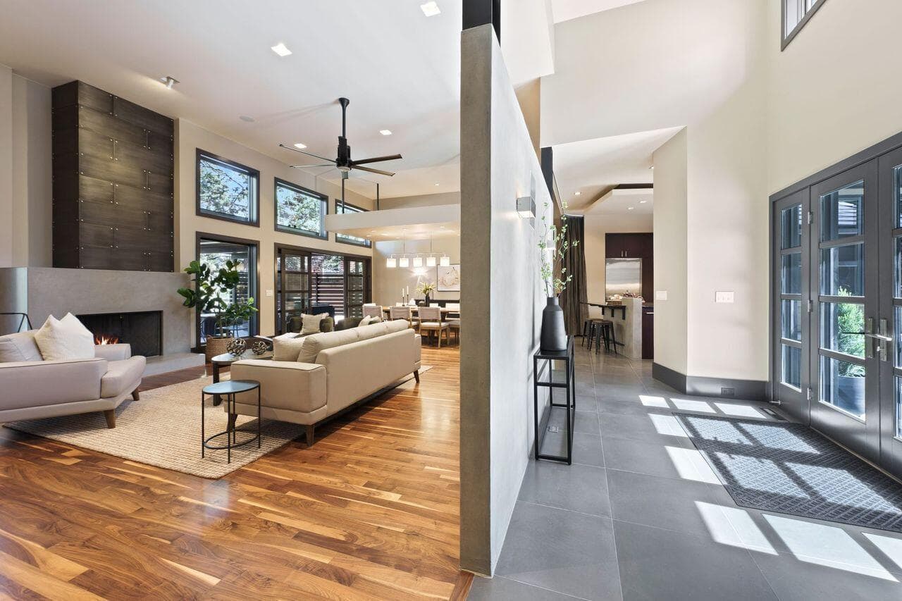 Main living space showcasing the home’s open yet thoughtfully defined layout, anchored by solid walnut flooring and a striking custom concrete feature wall that creates separation while maintaining a sense of openness. Expansive windows and high ceilings fill the space with natural light, while the great room flows seamlessly into the dining area and kitchen beyond. A modern fireplace, custom millwork, and integrated design elements reflect the home’s exceptional level of craftsmanship and cohesive warm modern aesthetic.