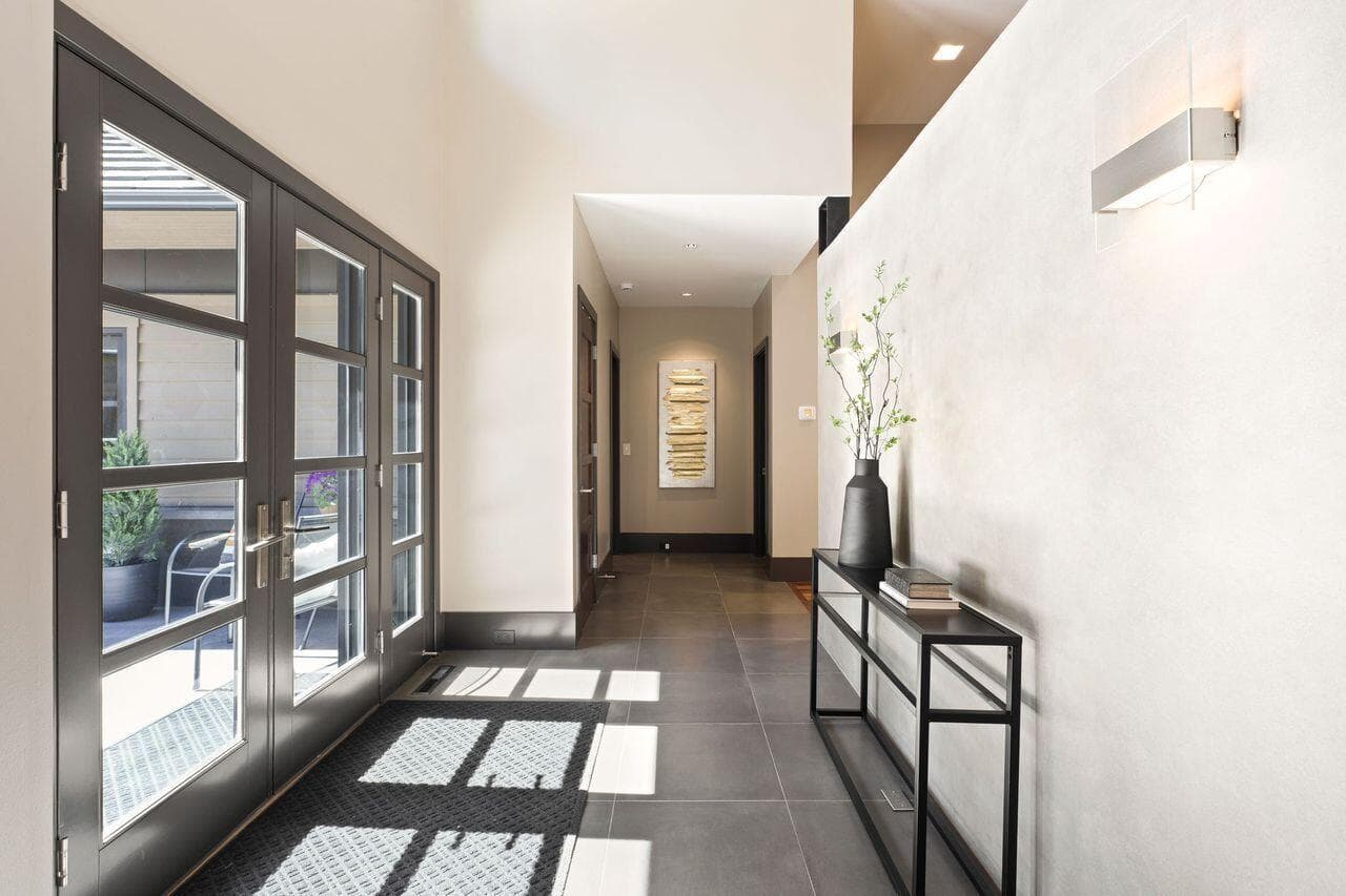 Entry corridor highlighting the home’s clean architectural lines and refined material palette, with large-format tile flooring and a continuation of the custom concrete feature wall. Natural light pours in through glass-paneled doors, creating a bright and welcoming transition from the private courtyard into the main living spaces. Thoughtful design details and art placement enhance the sense of calm, modern simplicity carried throughout the home.