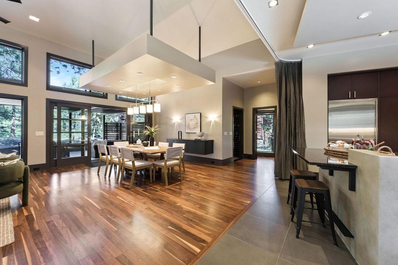 Dining area positioned at the heart of the home, highlighted by rich solid walnut flooring, soaring ceilings, and a striking suspended architectural element that adds both definition and visual interest. Floor-to-ceiling windows and glass doors frame views of the surrounding pines while filling the space with natural light. Subtle architectural features allow for flexibility between open living and more intimate entertaining, while maintaining a seamless connection to the kitchen and outdoor spaces.