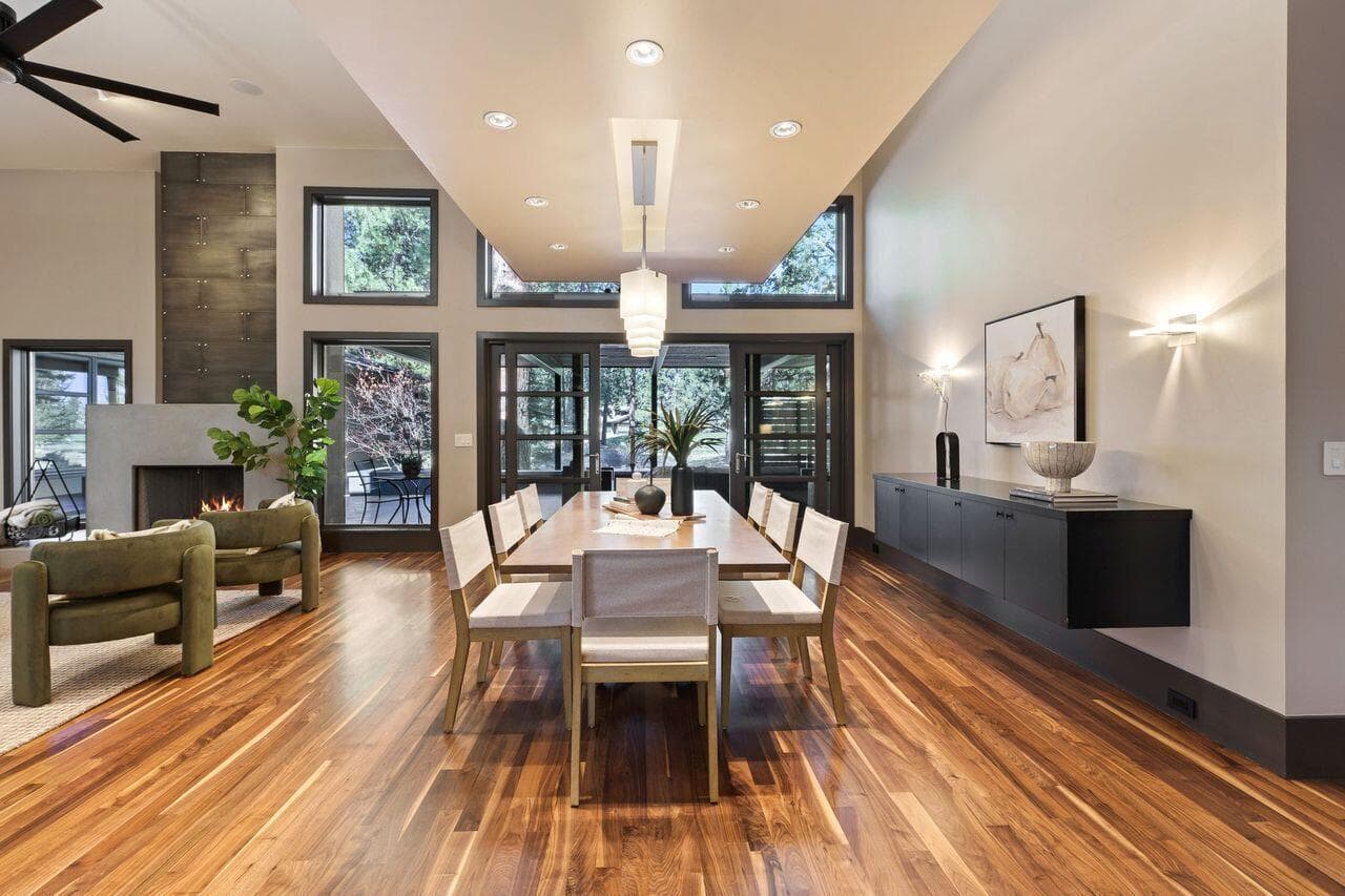 Formal dining space framed by rich solid walnut flooring and a dramatic ceiling detail that anchors the room while adding architectural depth. Expansive windows and glass doors invite natural light and create a seamless connection to the surrounding landscape and outdoor living areas. Thoughtfully designed with both scale and intimacy in mind, the space flows effortlessly into the adjacent living area, offering flexibility for everyday living or more refined entertaining.