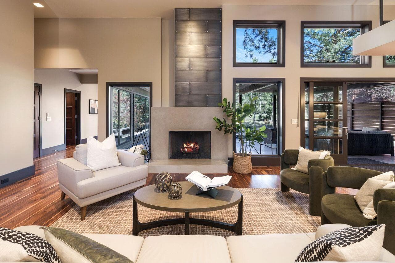 Great room centered around a modern fireplace with a custom concrete surround, complemented by vertical wood detailing and expansive windows that frame the surrounding pines. Warm walnut flooring and clean architectural lines create a comfortable, light-filled gathering space with a strong connection to the outdoors.