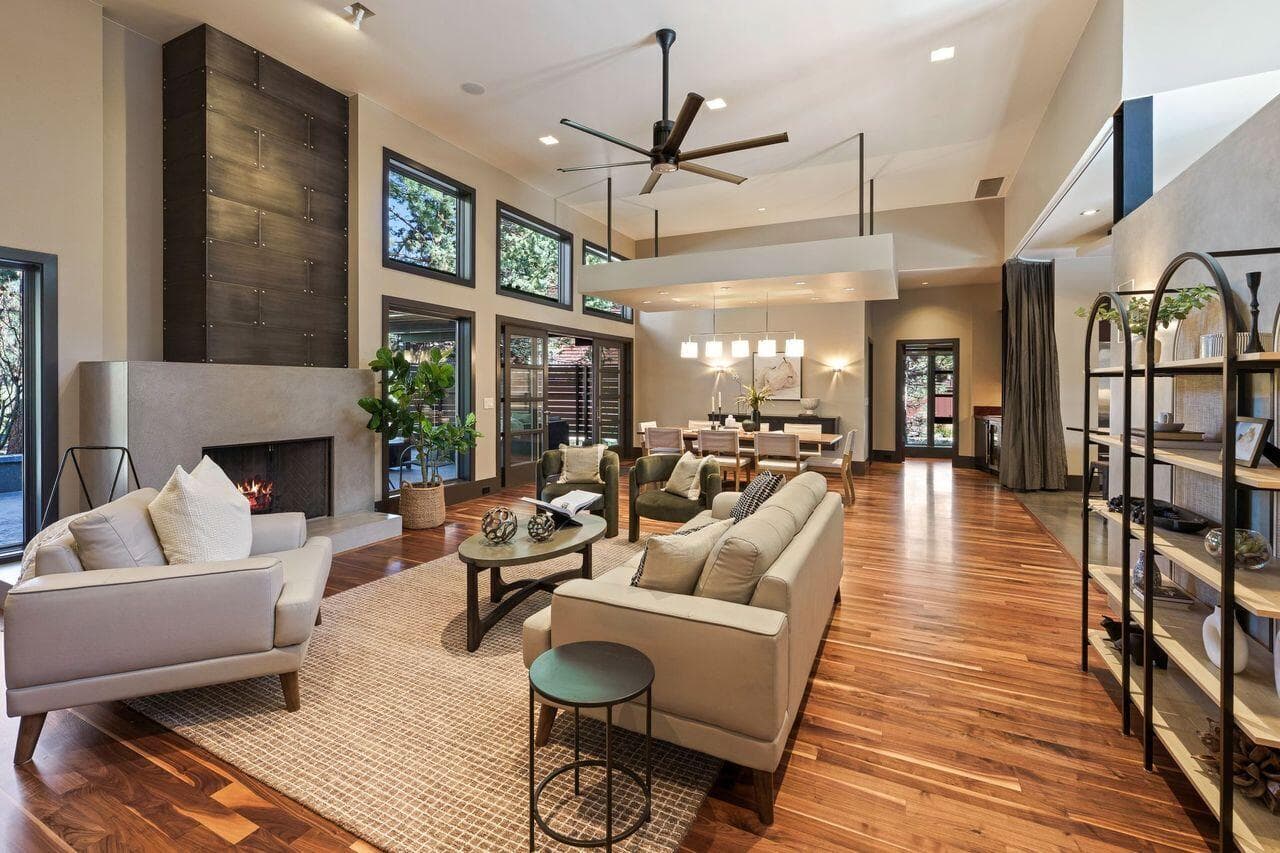 Open-concept great room and dining area featuring solid walnut flooring, a modern fireplace with custom concrete surround, and striking vertical wood detailing. Expansive windows bring in natural light and frame views of the surrounding pines, while the thoughtful layout offers both connection and separation between living spaces for comfortable everyday living and entertaining.