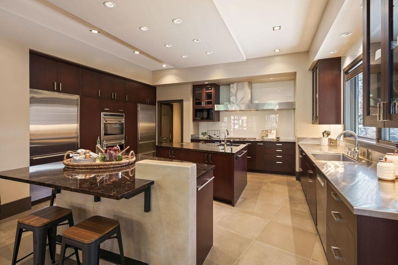 Well-appointed kitchen with custom cabinetry, stone countertops, and a large center island with seating, complemented by high-end appliances and a highly functional layout. Spandrel glass wall surfaces add a sleek, reflective quality while offering exceptional ease of cleaning—an elegant and practical design detail that enhances both form and function.