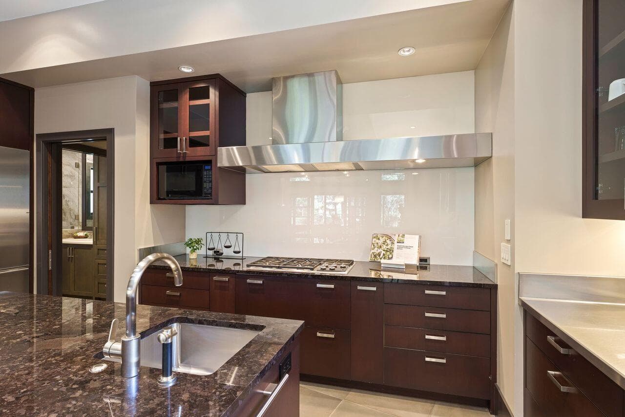 Kitchen workspace featuring a Wolf gas cooktop and microwave, stainless steel hood, stone countertops, and custom cabinetry, thoughtfully designed for both everyday use and more refined cooking. Spandrel glass wall surfaces introduce a sleek, reflective finish while offering exceptional ease of cleaning—an elegant balance of function and design.