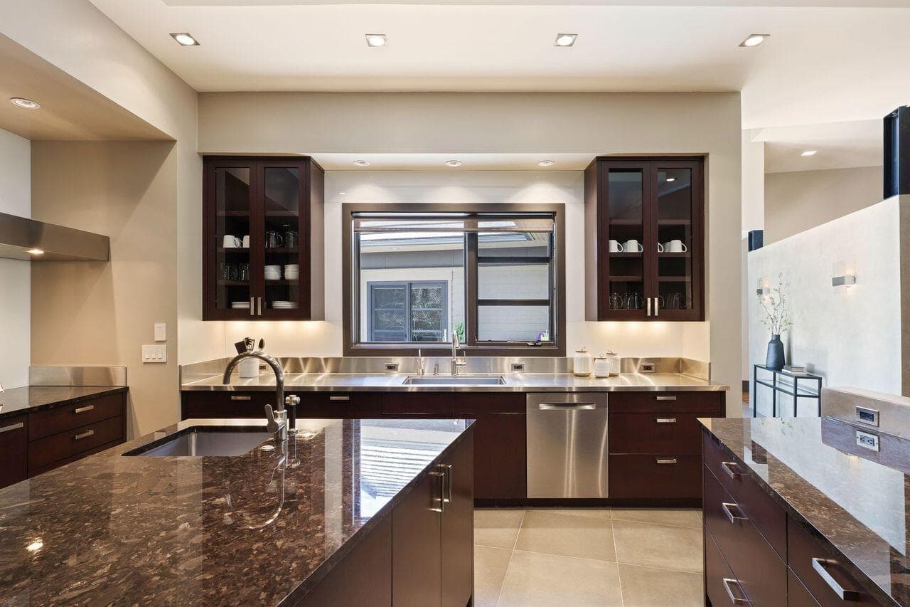 Thoughtfully designed kitchen with stainless steel countertops and sink, custom cabinetry, and a window over the sink bringing in natural light. Spandrel glass backsplash adds a sleek, reflective finish that enhances the space while offering exceptional ease of cleaning.