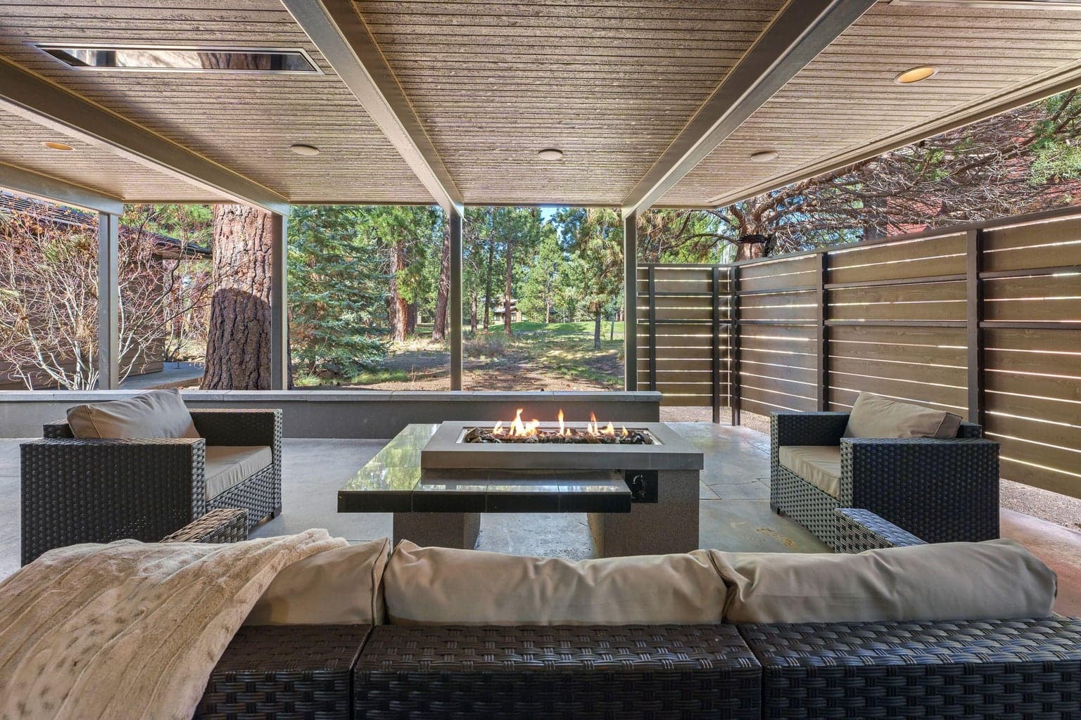 Covered outdoor living with a built-in concrete fire feature, recessed heaters, and privacy screening—perfect for year-round entertaining with a peaceful, treed backdrop.