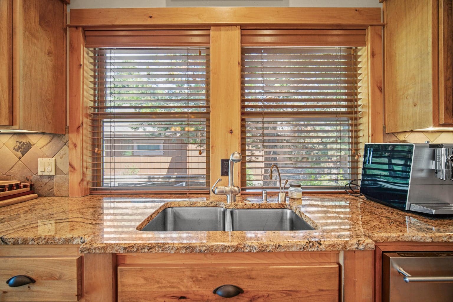 Kitchen sink framed by wood-trimmed windows, granite counters, tile backsplash, and warm natural cabinetry.