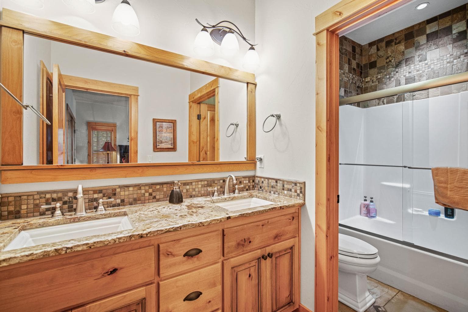Dual vanity en-suite bath with granite counters, wood cabinetry, framed mirror, and tile-accented tub and shower area.