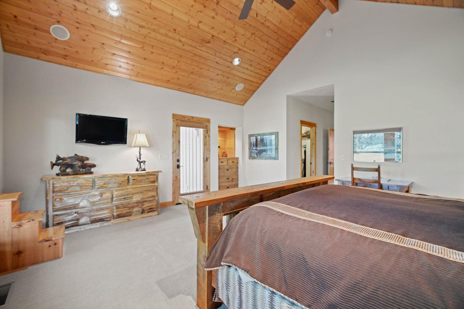Spacious main-level primary suite positioned on one side of the home, offering a quiet retreat with warm wood trim, large windows, and comfortable separation from the central gathering spaces.