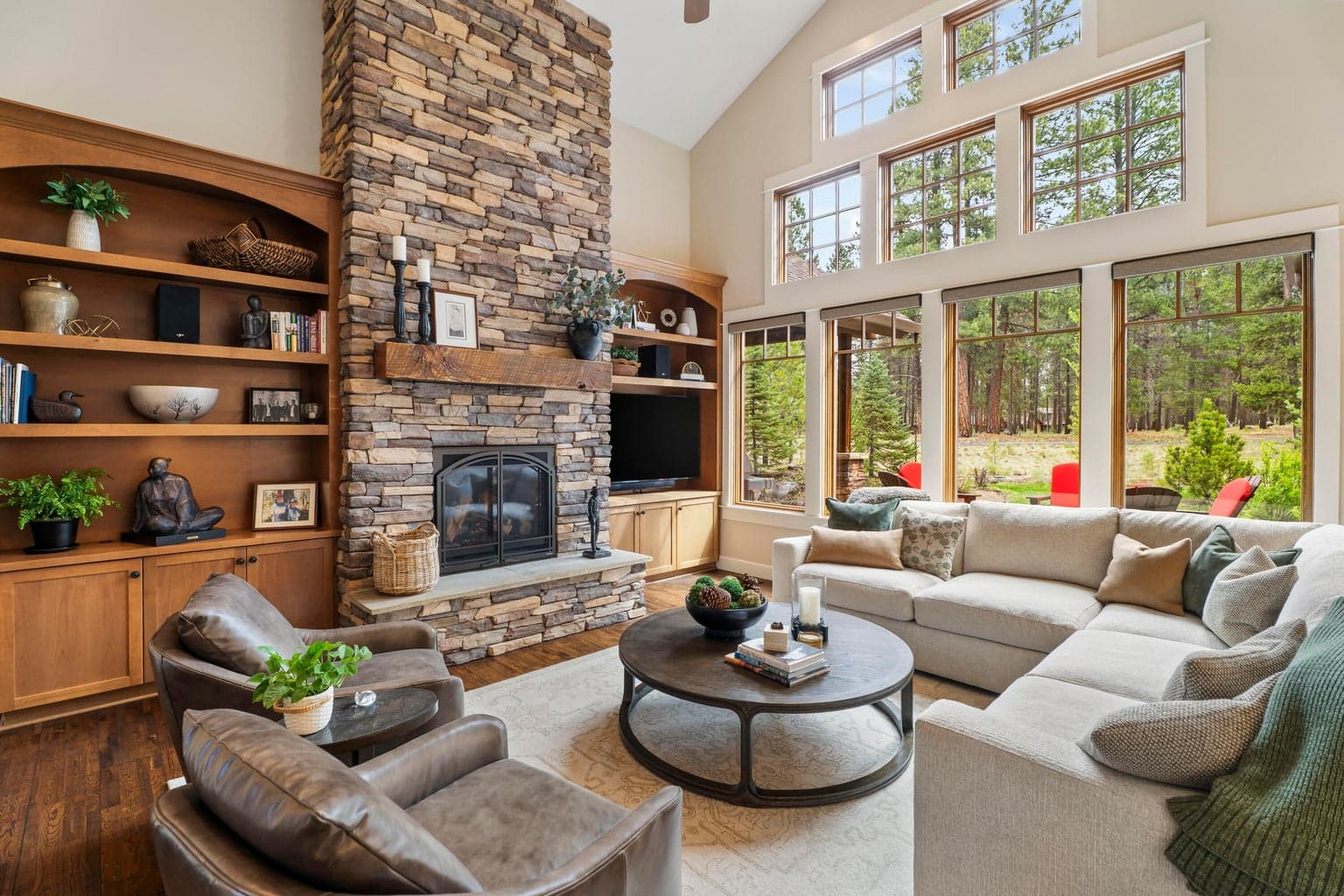 Soaring ceilings, a dramatic floor-to-ceiling stone fireplace, and expansive windows create a warm and light-filled great room overlooking the natural landscape.