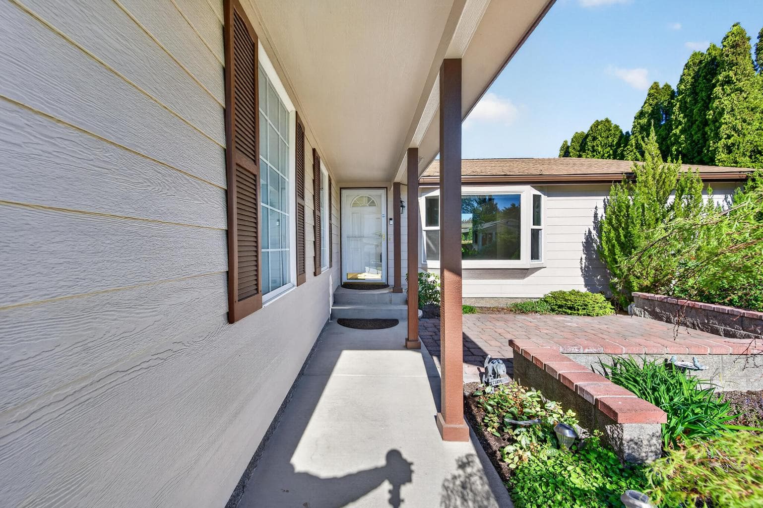 Extended covered walkway, front paver patio bordered by flower beds and landscaping.