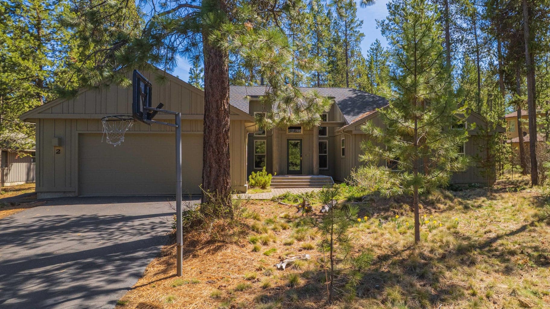 Front exterior view showing the home’s North Sunriver setting, paved driveway, and lightly treed natural landscape.