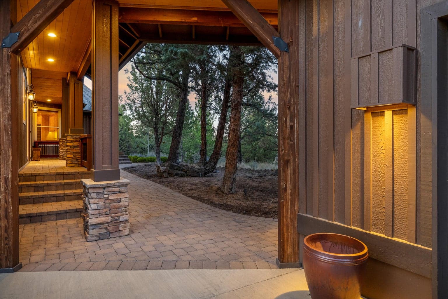 A covered paver pathway leads to the home’s thoughtfully positioned entry, where the approach unfolds through mature trees and warm architectural details before opening inside to the landscaped grounds and Cascade Mountain views beyond.