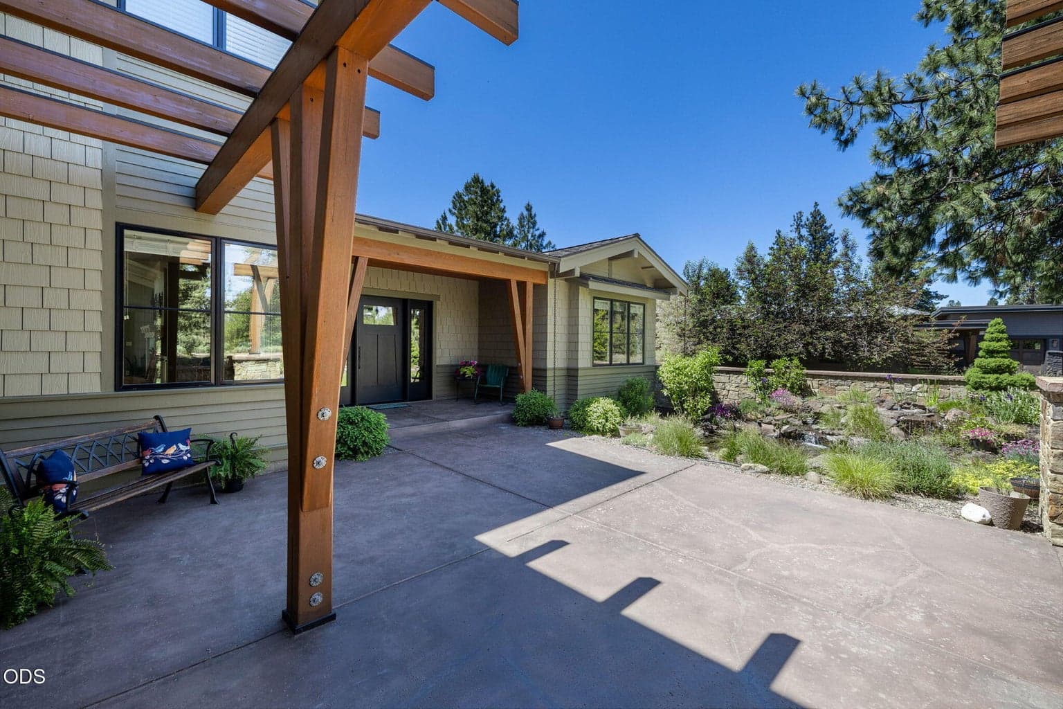 Expansive front courtyard patio framed by timber beams, stone walls, mature landscaping, and peaceful water feature views.