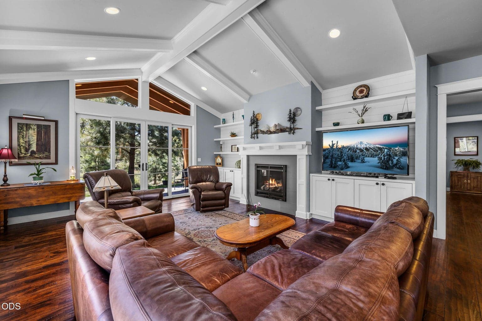 Vaulted great room with fireplace, built-ins, French doors, and expansive windows framing the surrounding trees and outdoor living area.