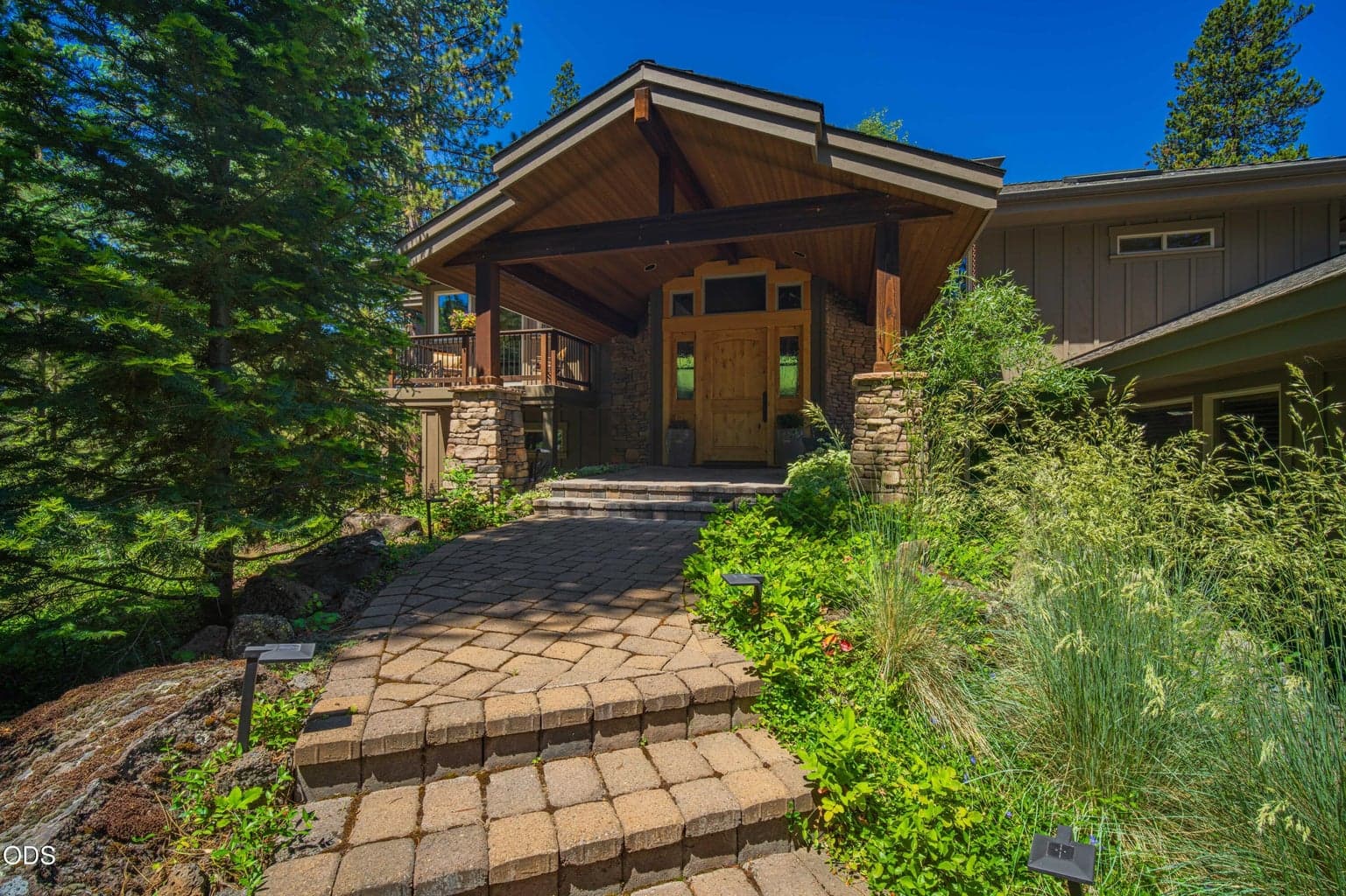 Close view of the timber-framed covered entry with substantial stone columns, wood front door, and carefully maintained landscaping around the home.