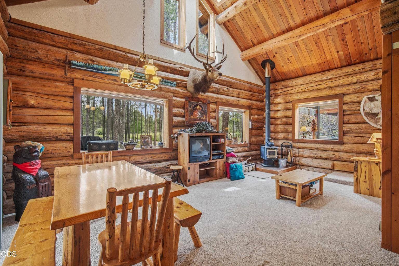 The vaulted great room brings together log construction, tall windows, warm wood ceilings, and a wood stove at the heart of the cabin.