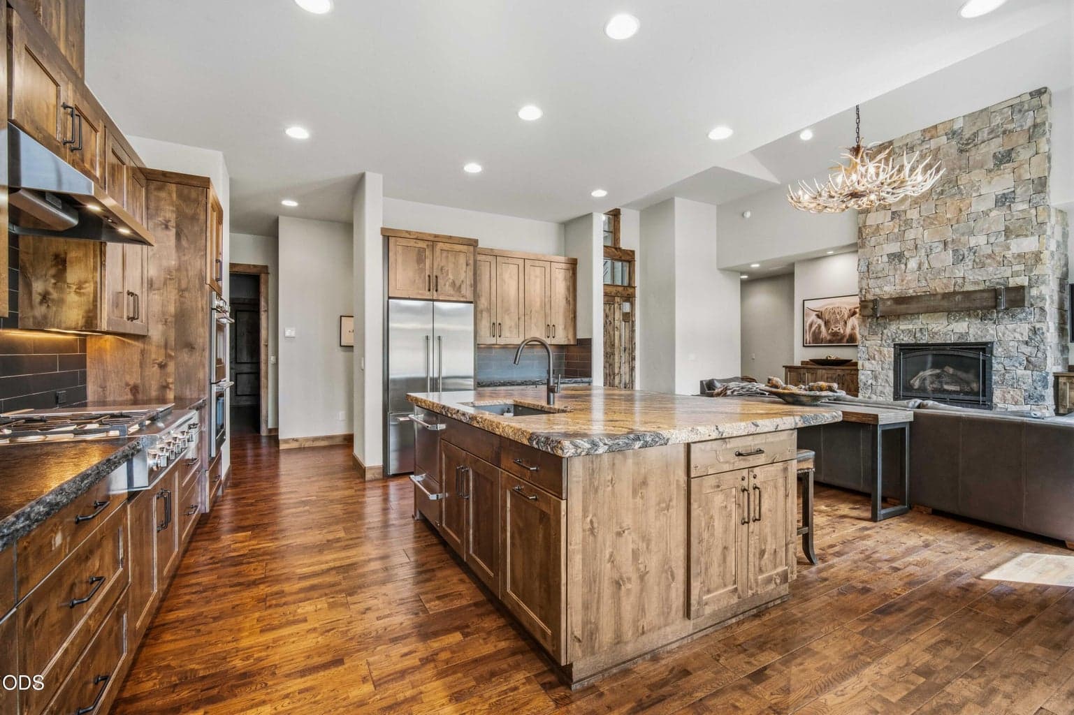 This view captures the home’s seamless indoor connection, from the kitchen to the great room and fireplace, with natural textures and refined finishes carried throughout.