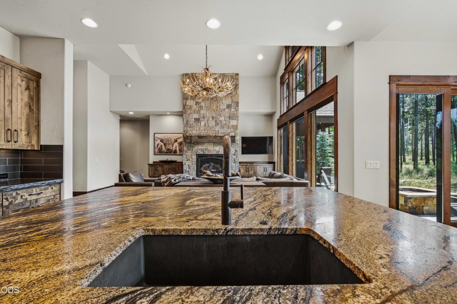 From the kitchen sink, the eye moves across richly grained counters to the stone fireplace and out toward the trees, a reminder that every space was designed to embrace the setting.