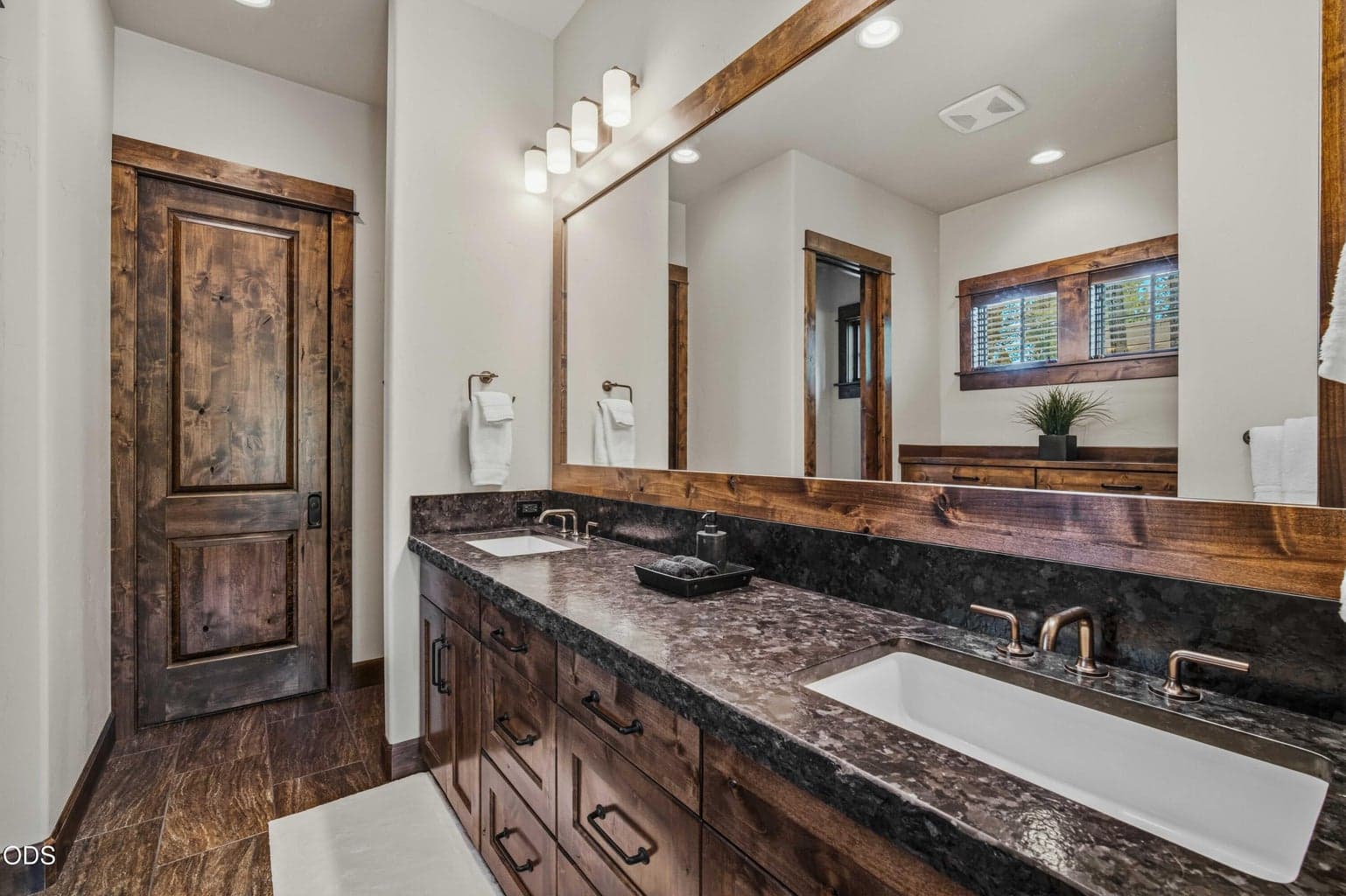 The primary bath offers a refined, spa-like feel with dual sinks, richly toned cabinetry, expansive mirror, and a warm material palette.