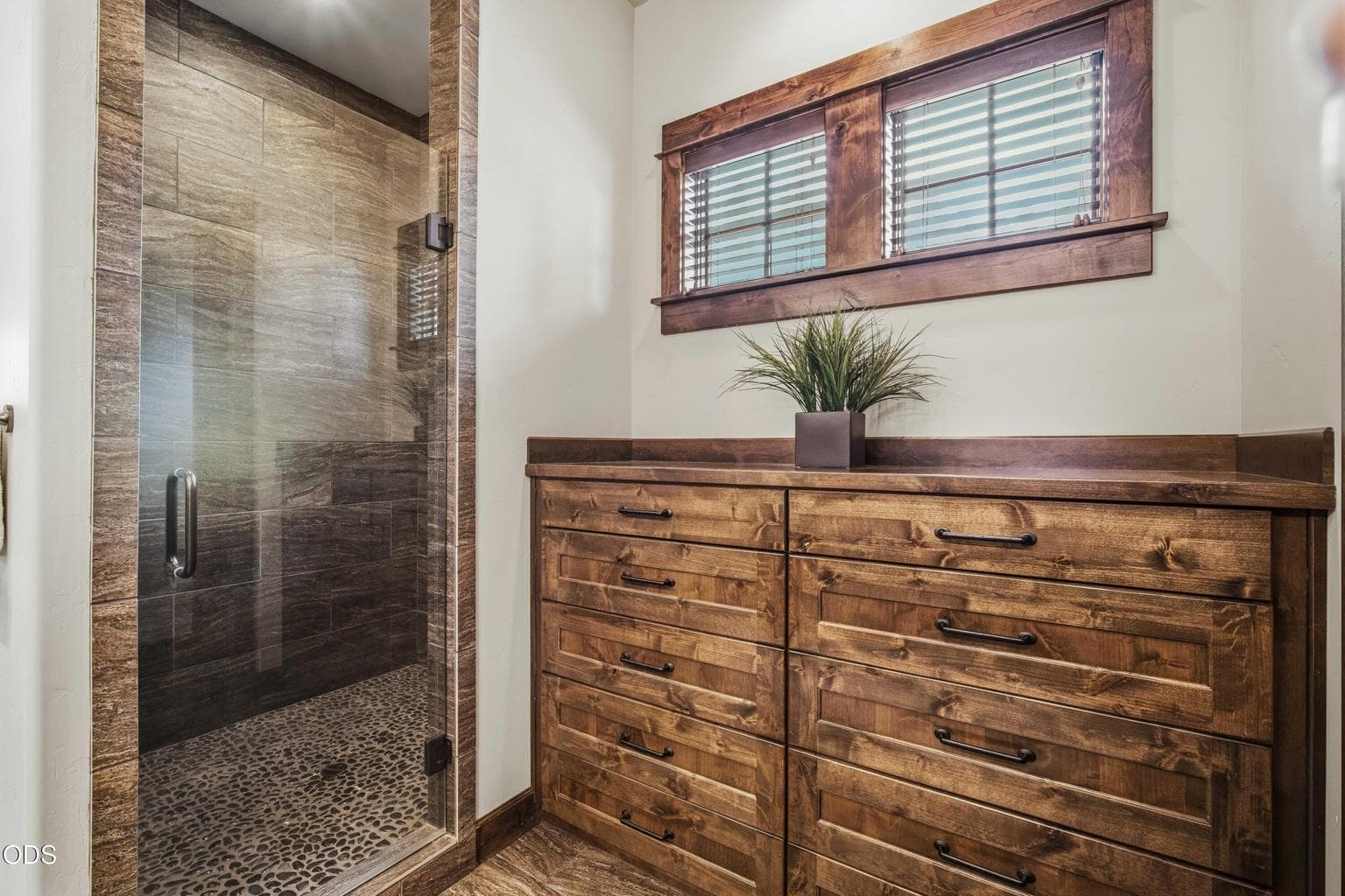 A glass-enclosed shower, pebble-style flooring, generous built-in storage, and warm wood cabinetry create a primary bath that feels both luxurious and grounded.