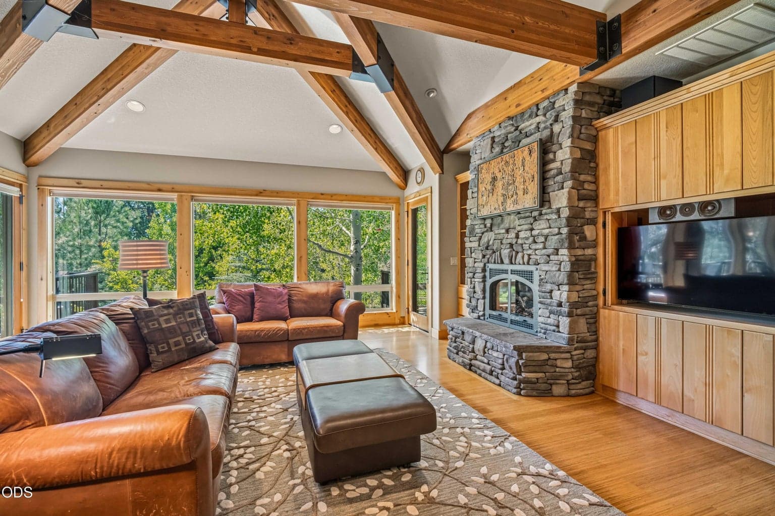 A substantial stone fireplace anchors the great room, accompanied by integrated cabinetry and media storage. Expansive windows preserve the home’s strong visual connection to the surrounding trees.