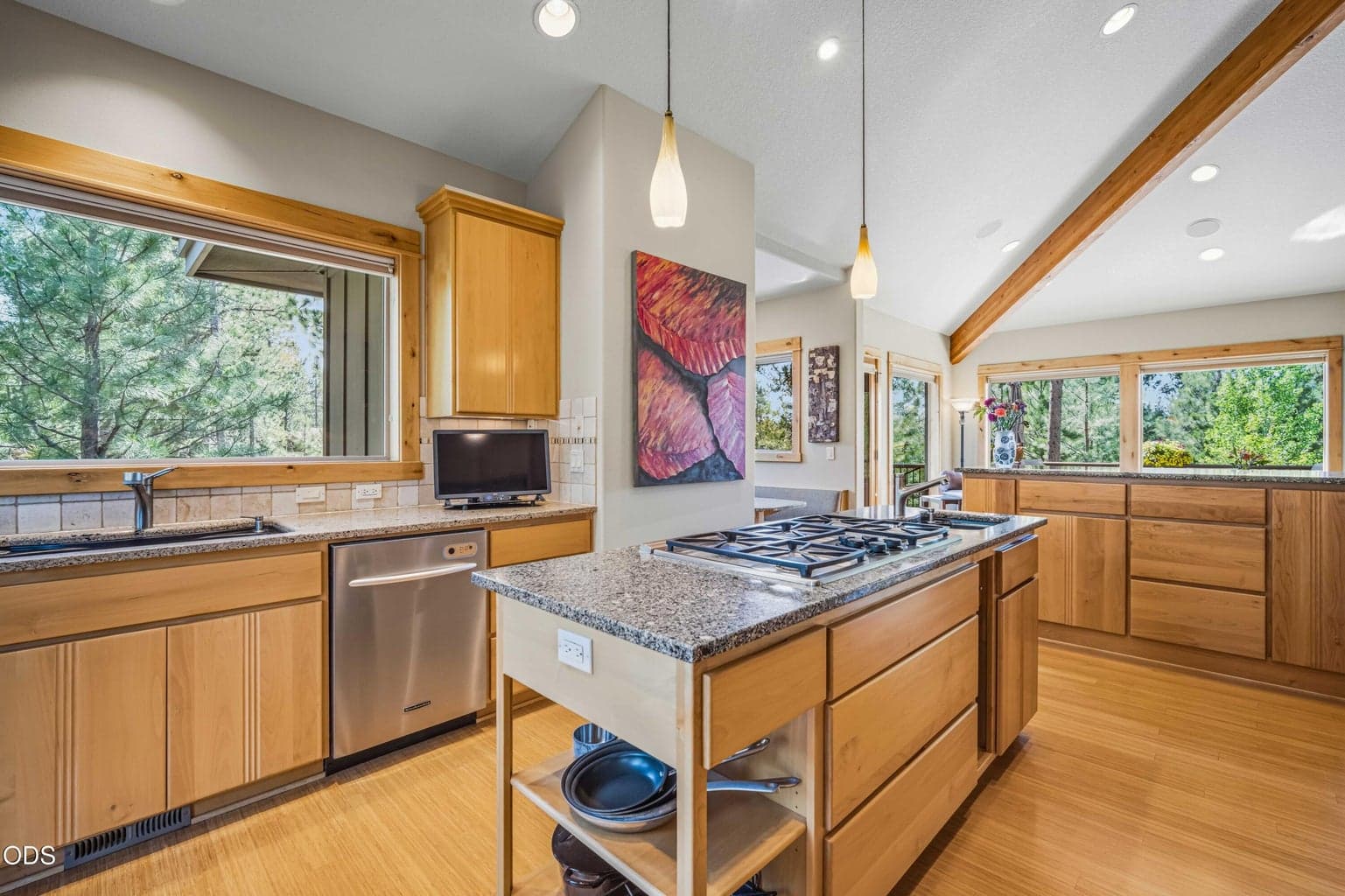 This expansive kitchen view highlights the central gas cooktop island, main sink beneath a broad forest-facing window, abundant natural wood cabinetry and vaulted ceiling with exposed beams. The open layout maintains a strong connection to the dining area and surrounding trees, supporting both everyday living and larger gatherings.