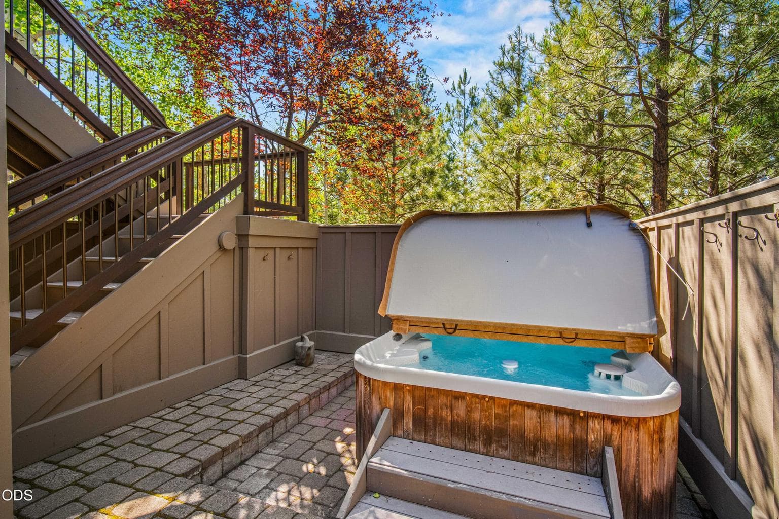 The hot tub occupies its own sheltered lower patio, offering privacy beneath the upper decks and a peaceful forest backdrop for year-round relaxation.