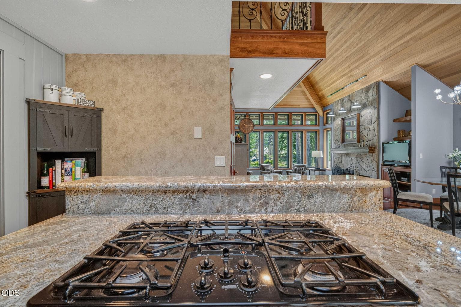 Granite island with gas cooktop overlooks open living space framed by wood beams and stone fireplace.