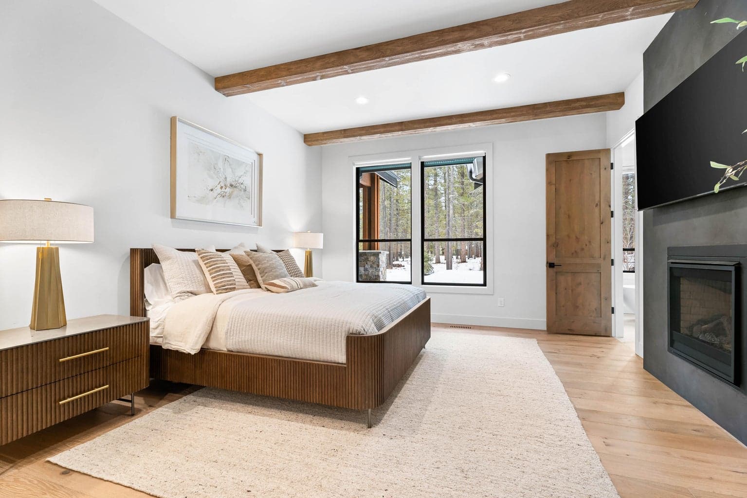 Spacious primary bedroom featuring warm wood flooring, exposed ceiling beams, a contemporary gas fireplace, and large windows framing the peaceful forested setting. A private en-suite bath is conveniently located just beyond. Photos are of a similar home previously built by the seller and are for illustrative purposes only. Actual design, finishes, furnishings and features may vary.