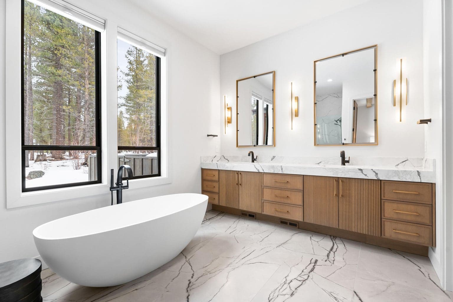 Spa-inspired primary bathroom featuring heated tile floors, a freestanding soaking tub, dual vanities with custom cabinetry, designer lighting, and oversized windows overlooking the forested setting. Photos are of a similar home previously built by the seller and are for illustrative purposes only. Actual design, finishes, furnishings and features may vary.