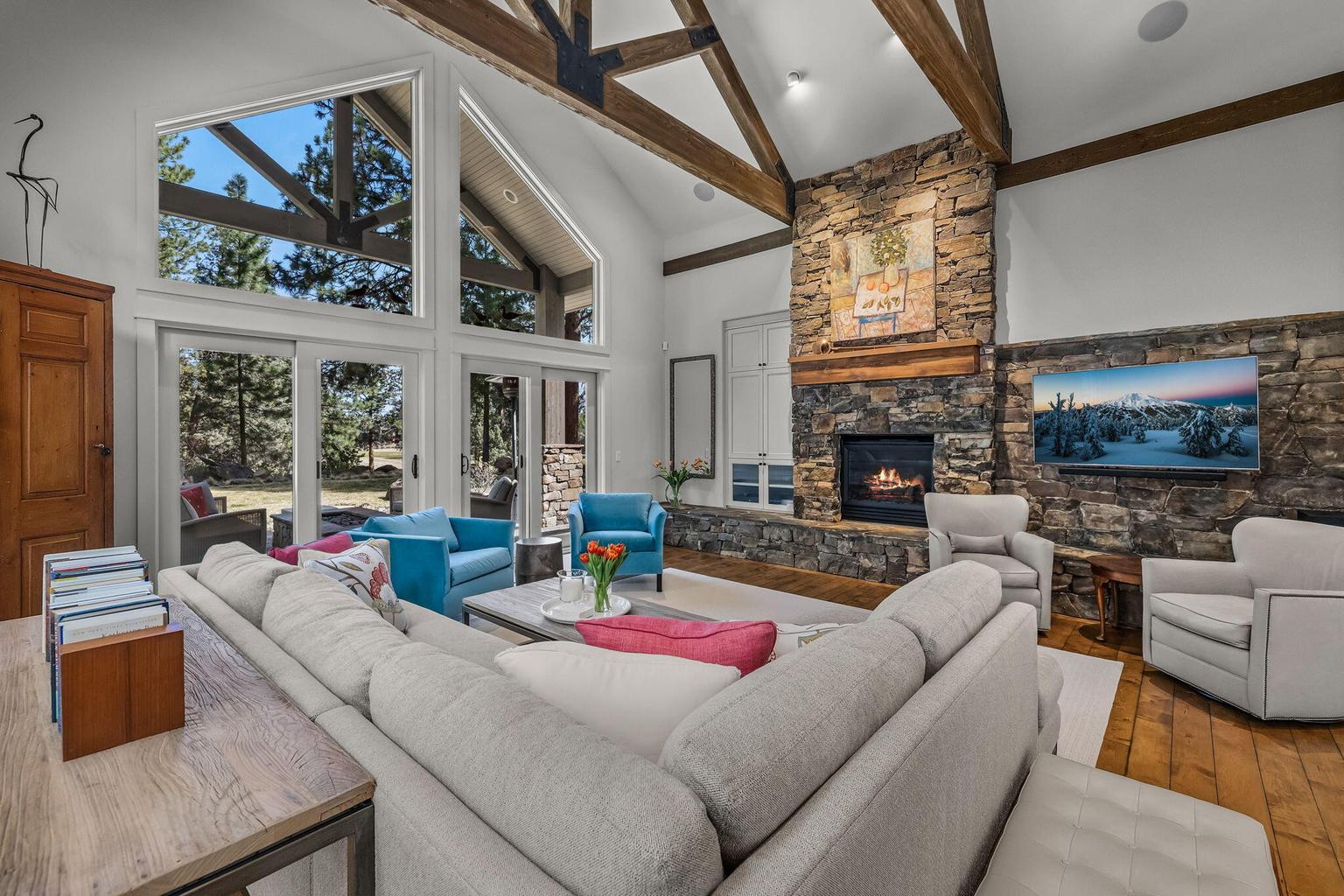 Walls of glass frame serene pine and golf course views, while the stone fireplace anchors the space and connects seamlessly to outdoor living beyond.