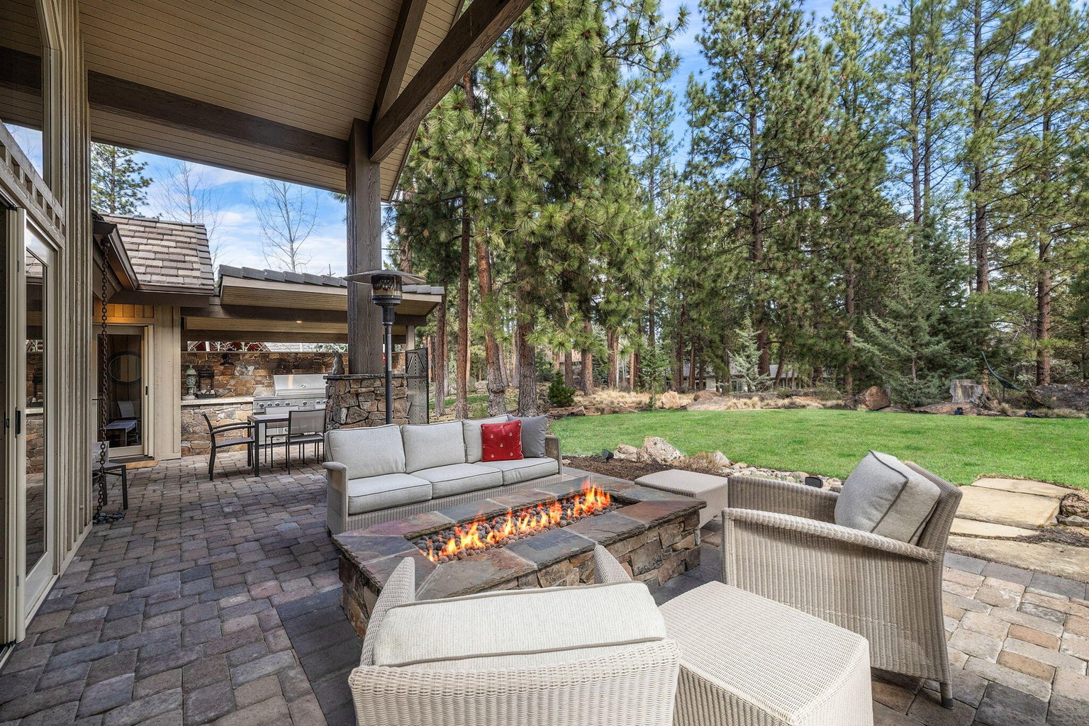 Expansive outdoor living set against a backdrop of pines, with covered patio, built-in kitchen, and multiple seating areas designed for relaxed entertaining.