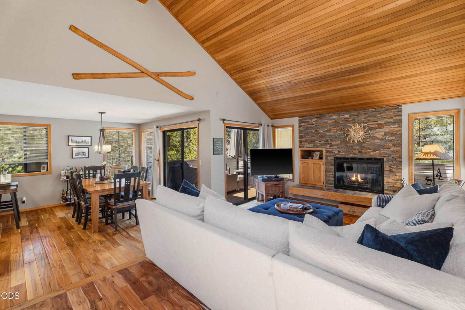 Bright and inviting great room with vaulted wood ceilings, hand-scraped Acacia wood floors, and a floor-to-ceiling stacked stone fireplace surround. Glass sliding doors connect the living area to the upper patio/deck, creating an easy indoor-outdoor flow for relaxing, entertaining, and enjoying the Sunriver setting.