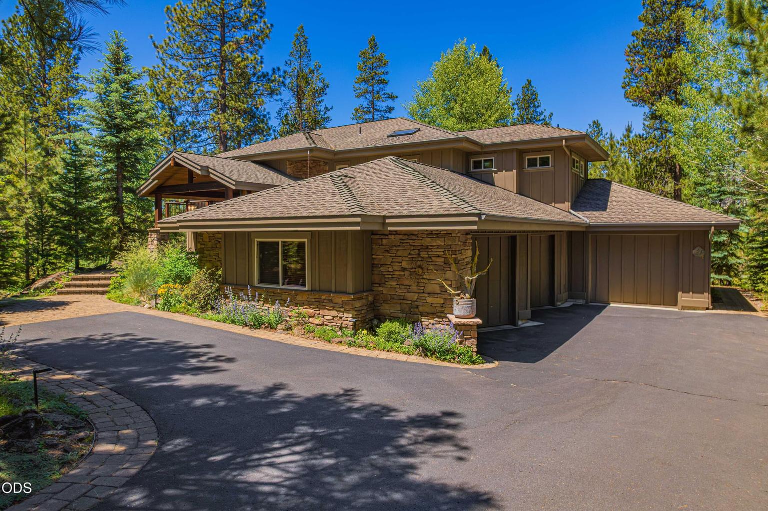 A broad exterior view highlights the Sunforest home’s substantial architecture, stone detailing, landscaped entry and generous three-car garage, all surrounded by the natural beauty of the Central Oregon forest.