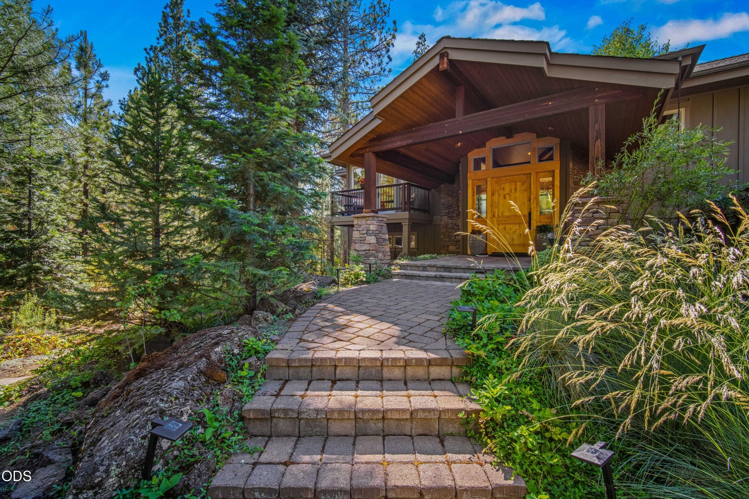 The covered entrance is framed by stone columns, warm wood detailing and mature trees, offering a sheltered and welcoming transition from the forested setting into the home.