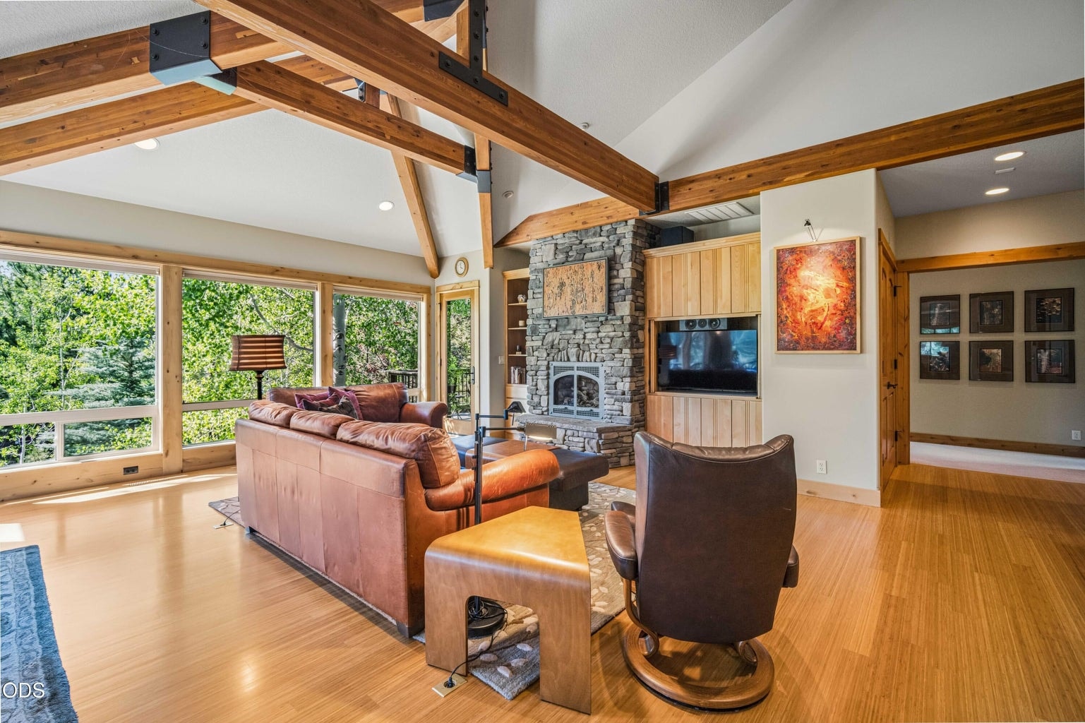 The great room combines architectural scale with warmth, including vaulted ceilings, exposed beams, a stone fireplace, built-ins and abundant natural light from windows overlooking the private forest setting.