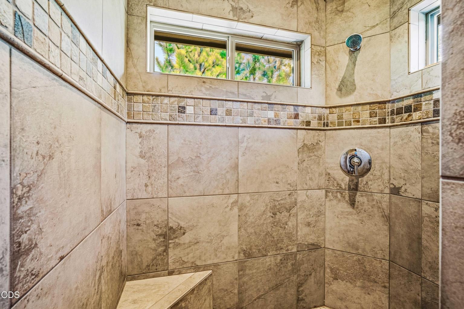 The primary bath includes a fully tiled walk-in shower with bench seating, natural light and a decorative mosaic accent complementing the home’s slate and stone finishes.