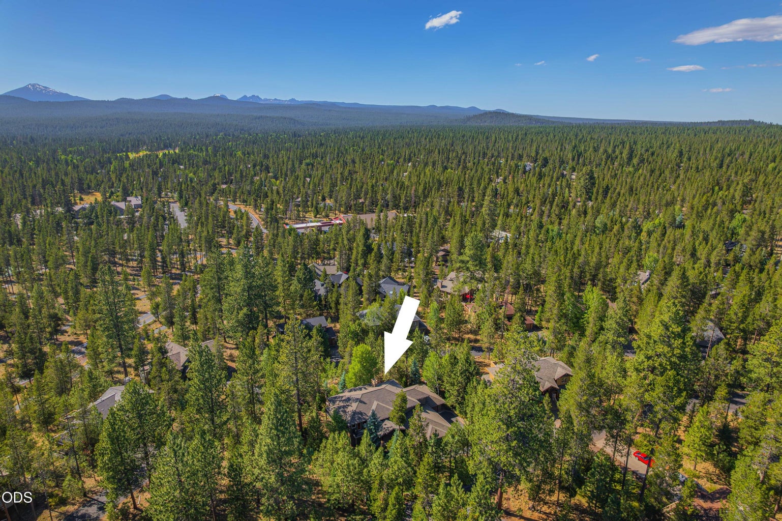 A dramatic aerial places 21 Wallowa Loop within Sunriver’s expansive forested landscape, with Mount Bachelor and the Central Oregon Cascades visible beyond. The arrow identifies the home and highlights its orientation toward the mountain views enjoyed from the dining area and upper decks.