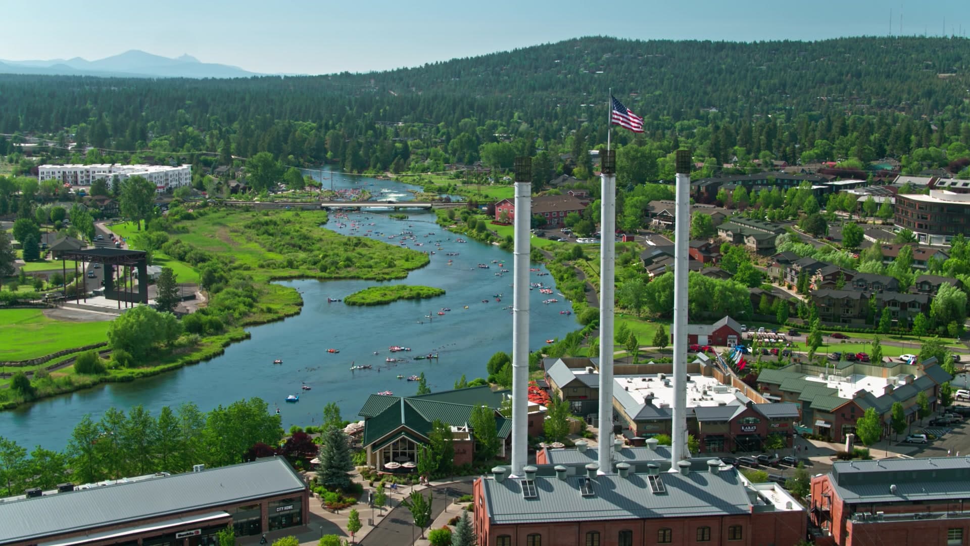 Old Mill District drone view with the American flag, the Deschutes River, and the Cascade mountains.